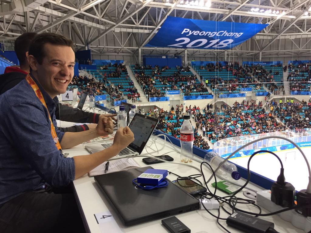Host and reporter, Anthony McLachlan sitting in press area. Looking at the camera he grips his fists with tension. Hundreds of fans in background with larger banner that reads, "PyeongChang 2018"