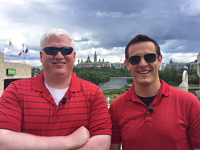 Dave Brown and Anthony McLachlan, wearing red shirts and sunglasses, stand together with Parliament Hill in the background.