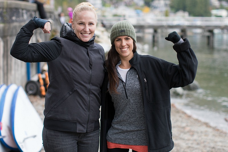Two women smile into the camera, flexing their arm muscles.