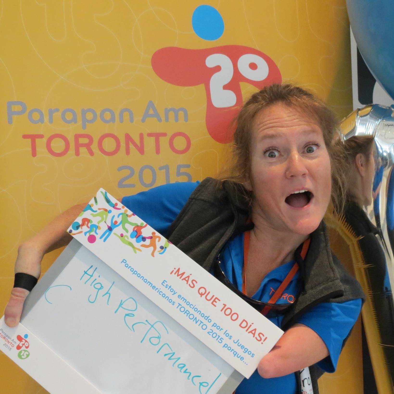 Tracy Schmitt holds her mouth open with an excited look on her face while standing in front of a banner for the Toronto 2015 Parapan Am Games. She’s holding a sign that reads, “High Performance.”