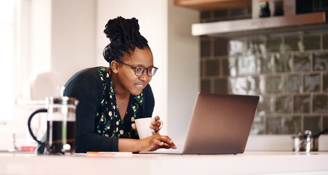 A woman sits at a table, looking at her laptop screen.