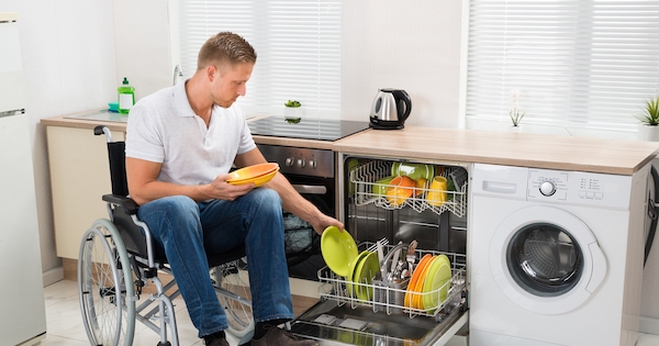 A man loads dishes into a dishwasher. He is using a wheelchair.