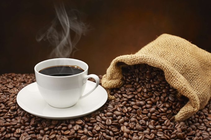A steaming cup of coffee sits on a heap of coffee beans spilling from a burlap sack. 