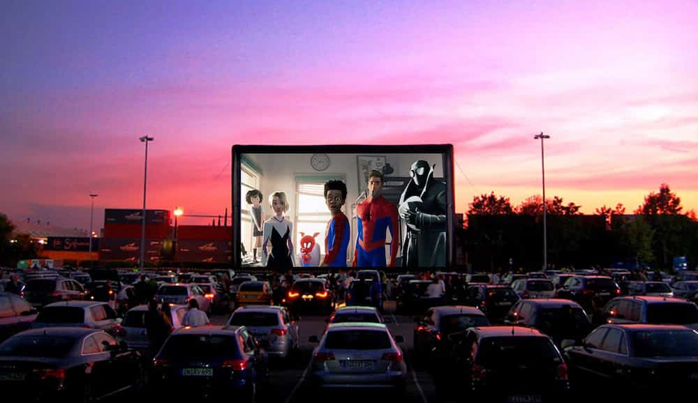 A drive-in theatre with several cars and Spiderman on screen with a sunset in the background.