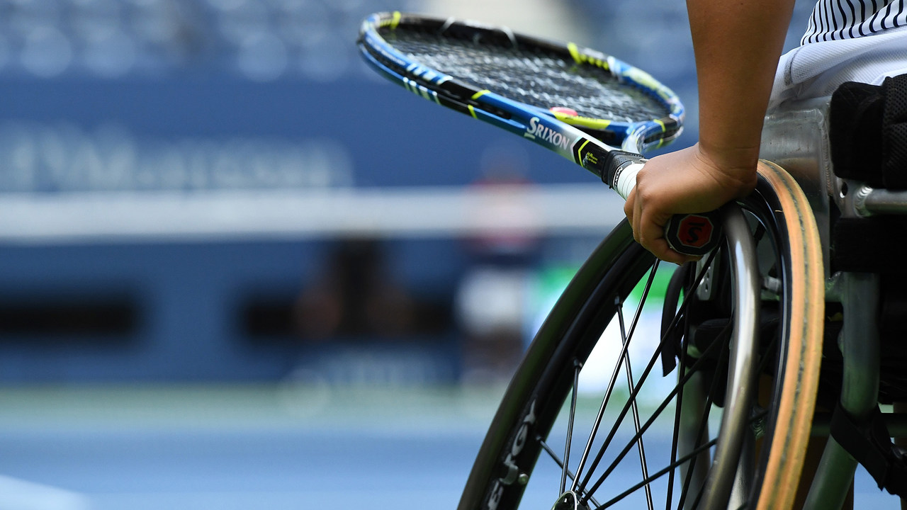 A closeup of someone's hand holidng a tennis racket and the wheel of their wheelchair.