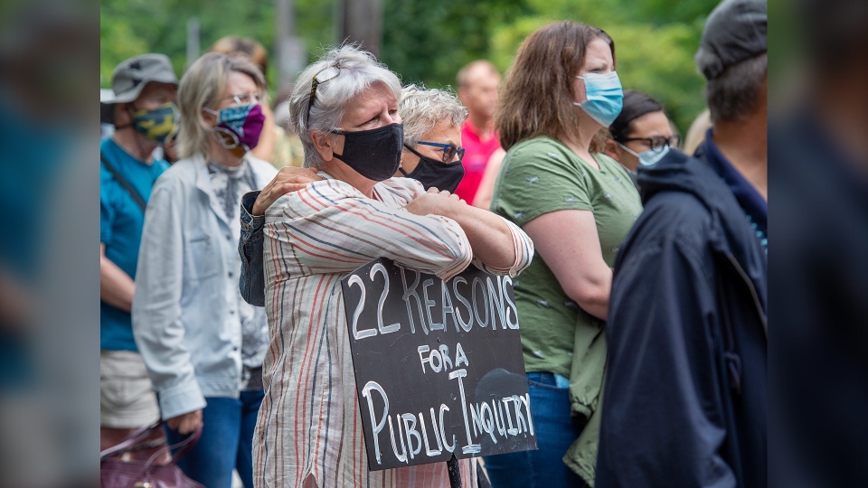 A woman with a mask stands alongside other protesters with her arms crossed and holding a sign that reads "22 reasons for a public inquiry"