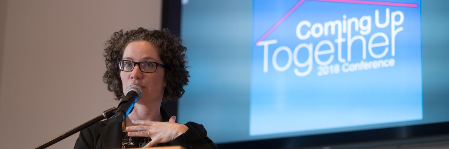Jacqueline Kennelly speaks at a podium. Behind her, a screen shows an abstract house and the words Coming Up Together 2018 Conference 