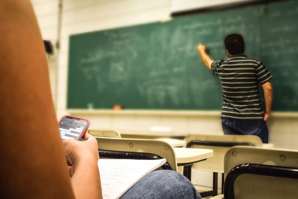 Picture shows one student writing on a chalkboard while a seated student examines her phone.   