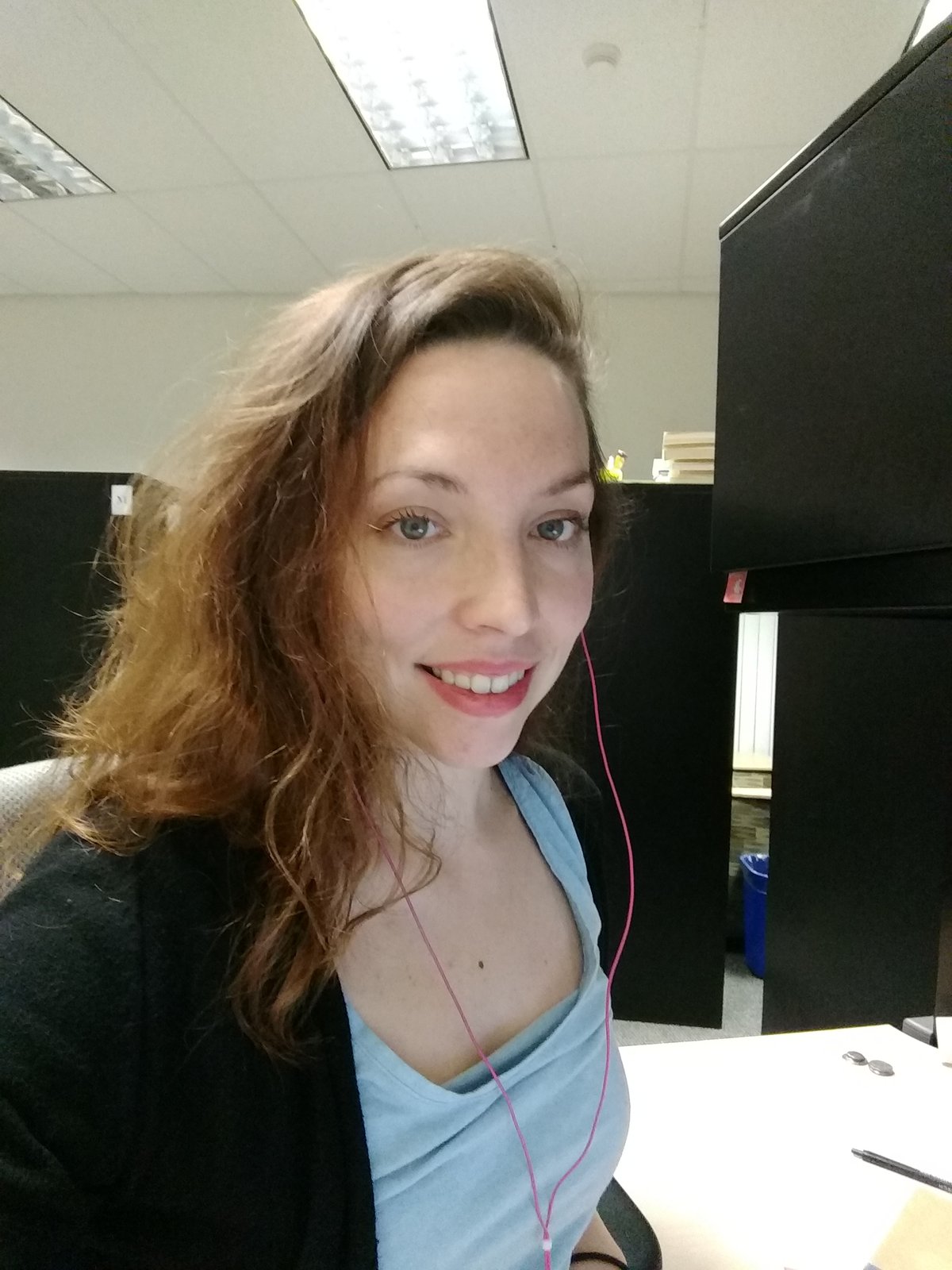 A seated brunette (Kristen Syme) wearing earbuds smiles into the camera.