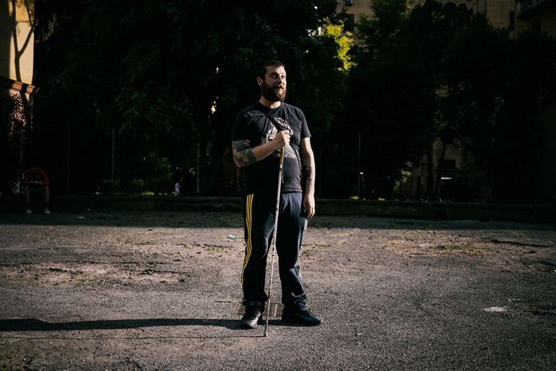 A bearded tattooed man standing in the middle of an empty lane leans on his white cane. 