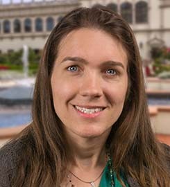 A woman (Suzanne Stolz) looks ahead and smiles. She has brown, shoulder length hair. The backdrop is an image of a building on the University of San Diego campus with a decorative pool in front of it.  