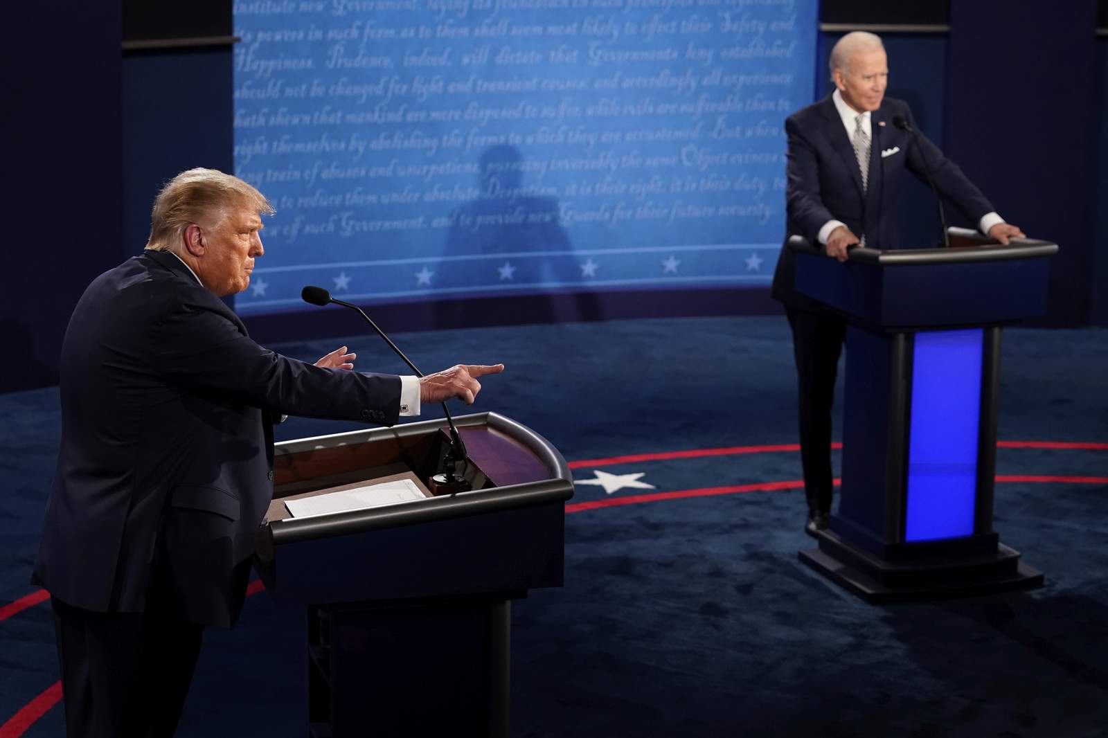 Donald Trump and Joe Biden stand behind their respective podiums during the recent US presidential debate. Trump is speaking and pointing while Biden is standing with his hands gripping the podium. They are both looking at the moderator who is outside of the frame. 