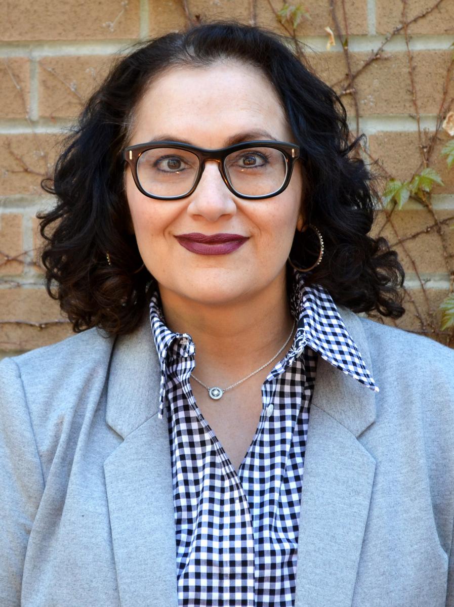 An Indo-Canadian woman (Ena Chadha) wearing black glasses, a powder blue blazer jacked and a checked, collared shirt looks at the camera, with a brick wall and branches in the background. 