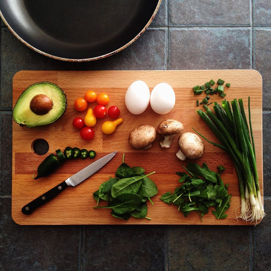 An avocado, mushrooms, eggs, green onions, a jalapeño and spinach are neatly organized on a wooden cutting board with knife sitting in the corner. 