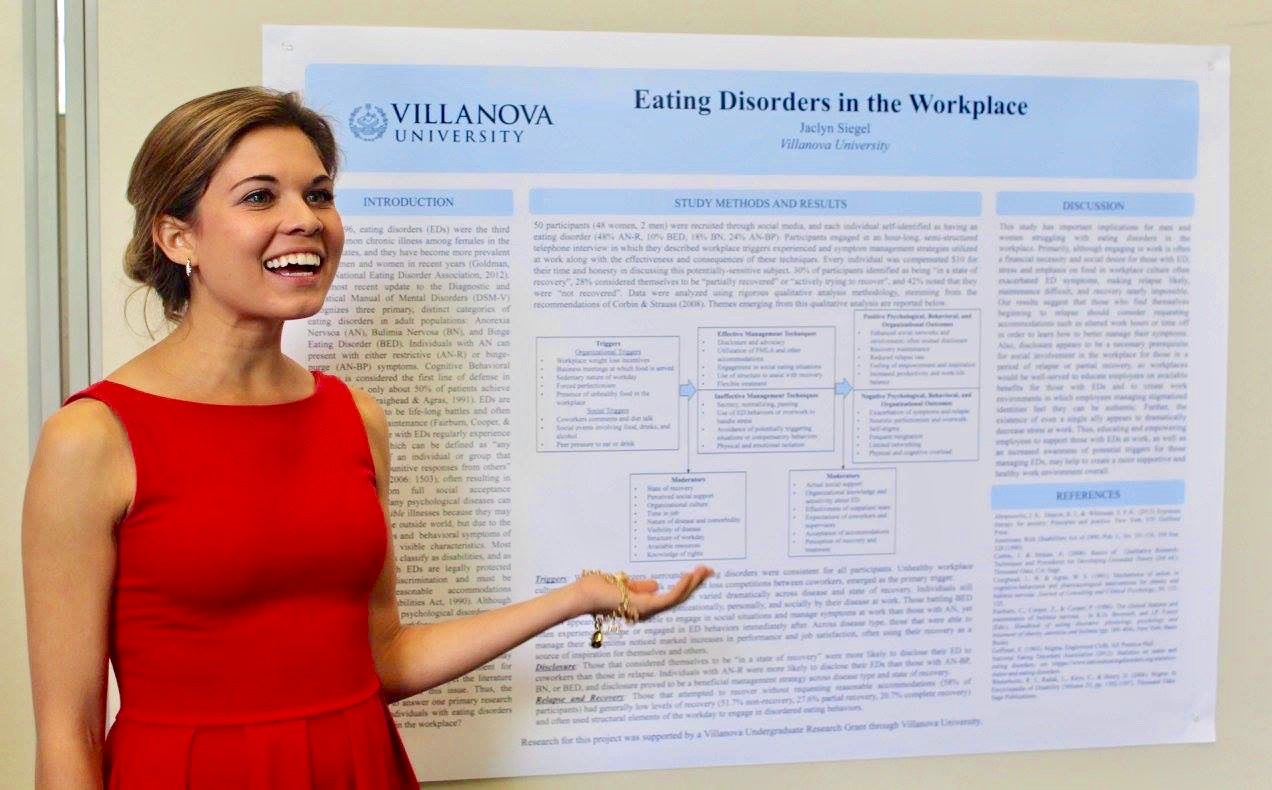 A smiling woman (Jaclyn Siegel) wearing a red dress gestures towards a wall chart titled “Eating Disorders in the Workplace”. 