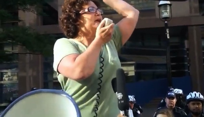 In a still taken from the documentary Judy vs Capitalism, Judy Rebick gives an impassioned speech to a rally in front of Toronto Police headquarters. Her left arm is raised to emphasise a point. Her right hand holds a microphone which is connected to a megaphone with a coiled wire. She is standing on a platform, which is outside of the frame. She stands above, and with her back to, several police officers who look on impassively.   