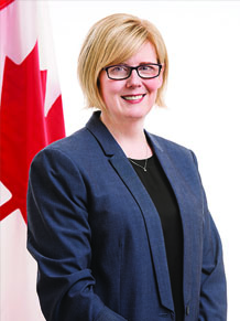 Carla Qualtrough smiles at the camera. A Canadian flag can be partially seen behind her right shoulder.