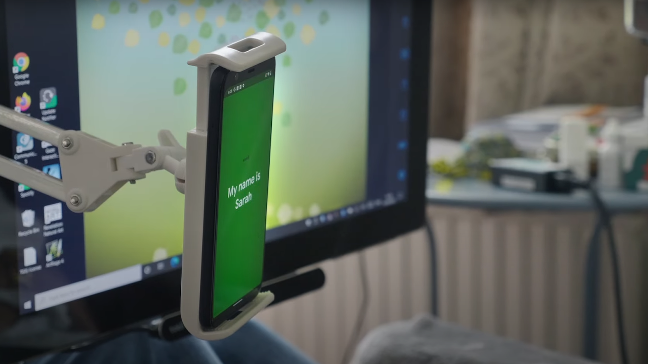 A smartphone held up on a phone holder with a green screen that reads "Hello my name is Sarah."