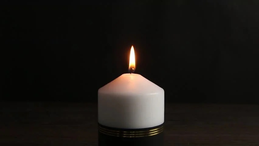 Image shows a short, tapered white candle with a black ribbon around its base, flickering against a black background. 