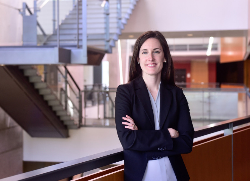 Image shows a brunette woman, Hilary Brown, smiling into the camera with her arms crossed. In the background is the lobby of a modern building. 