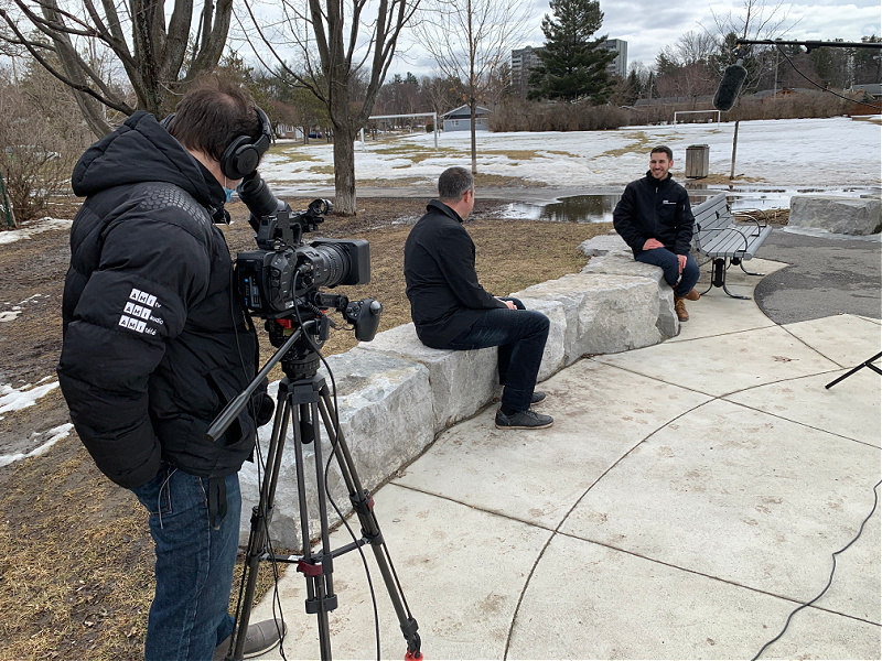 Two men are socially distanced during an interview in a park. A television camera films them.