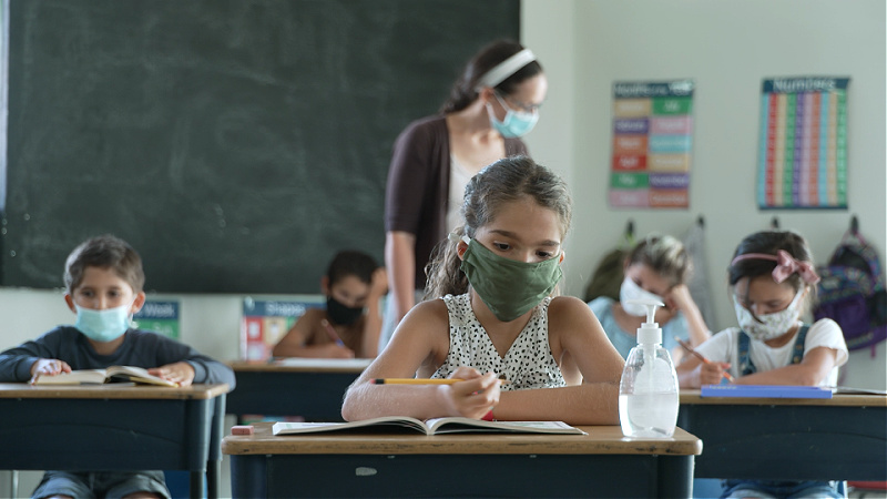 Children sit in a classroom, wearing surgical masks.