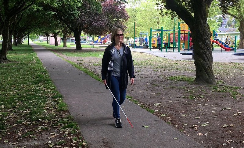 A woman, wearing sunglasses and dressed for cool weather, walks using a white cane.