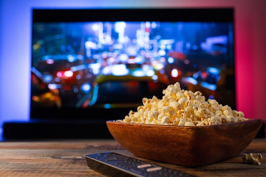 a bowl of popcorn is in focus with a television remote next to it. a movie plays in the background