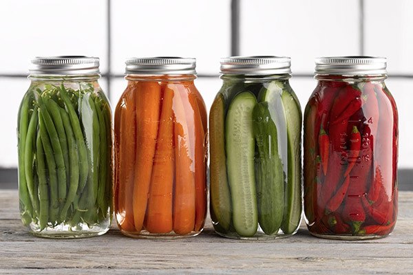 four jars of food are neatly sliced and packed into jars for preservation. From left to right, green stringy beans, beefy carrots, slices of cucumber and hot peppers