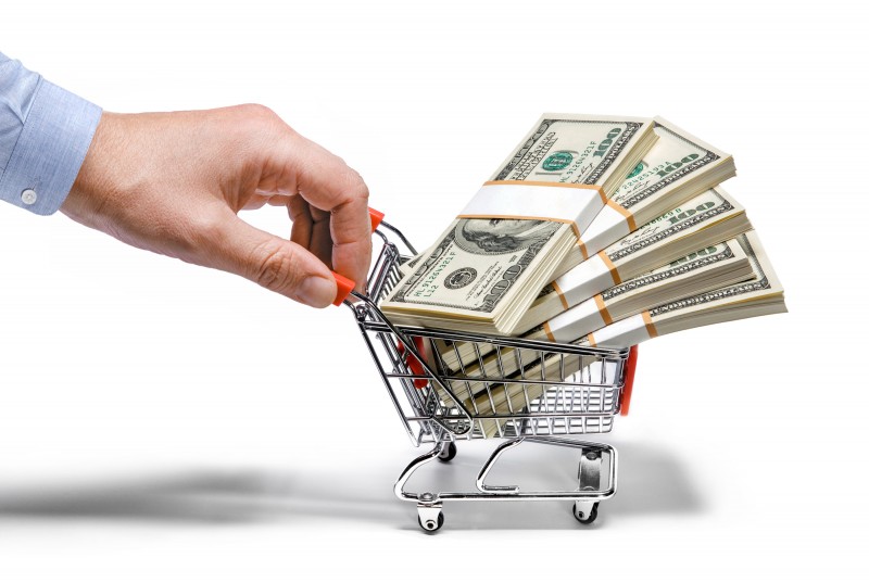 A hand is holding onto a miniature shopping cart. Stacks of cash are neatly piled inside the cart.