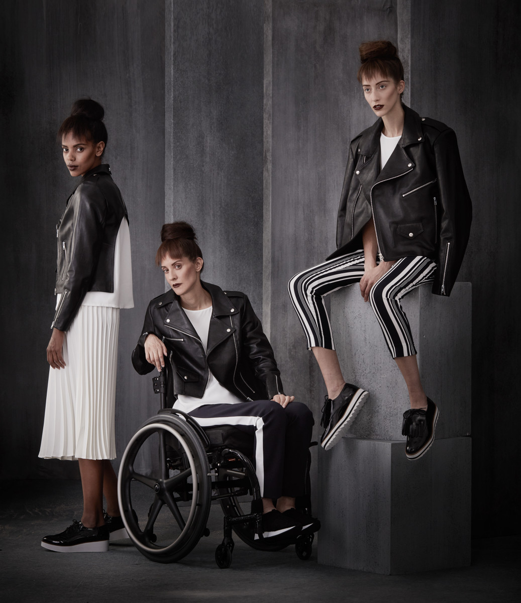 3 women are posing. From left to right, a woman stands side profile looking at the camera. The next model in a wheelchair arm rested in a relaxed pose. The last model sitting on two tall cubes. Stylish leather and casual wear worn by all three.