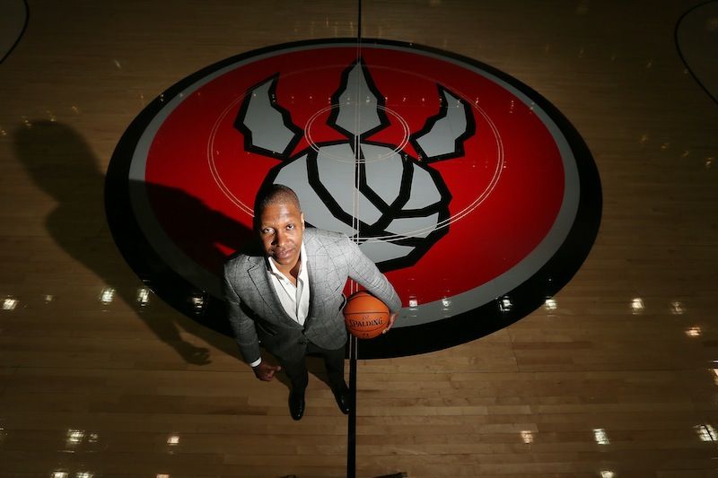 Masai Ujiri stands on a basketball court with a ball under his arm. it's a closeup birdseye view as he stands beside the raptors logo on the floor. 
