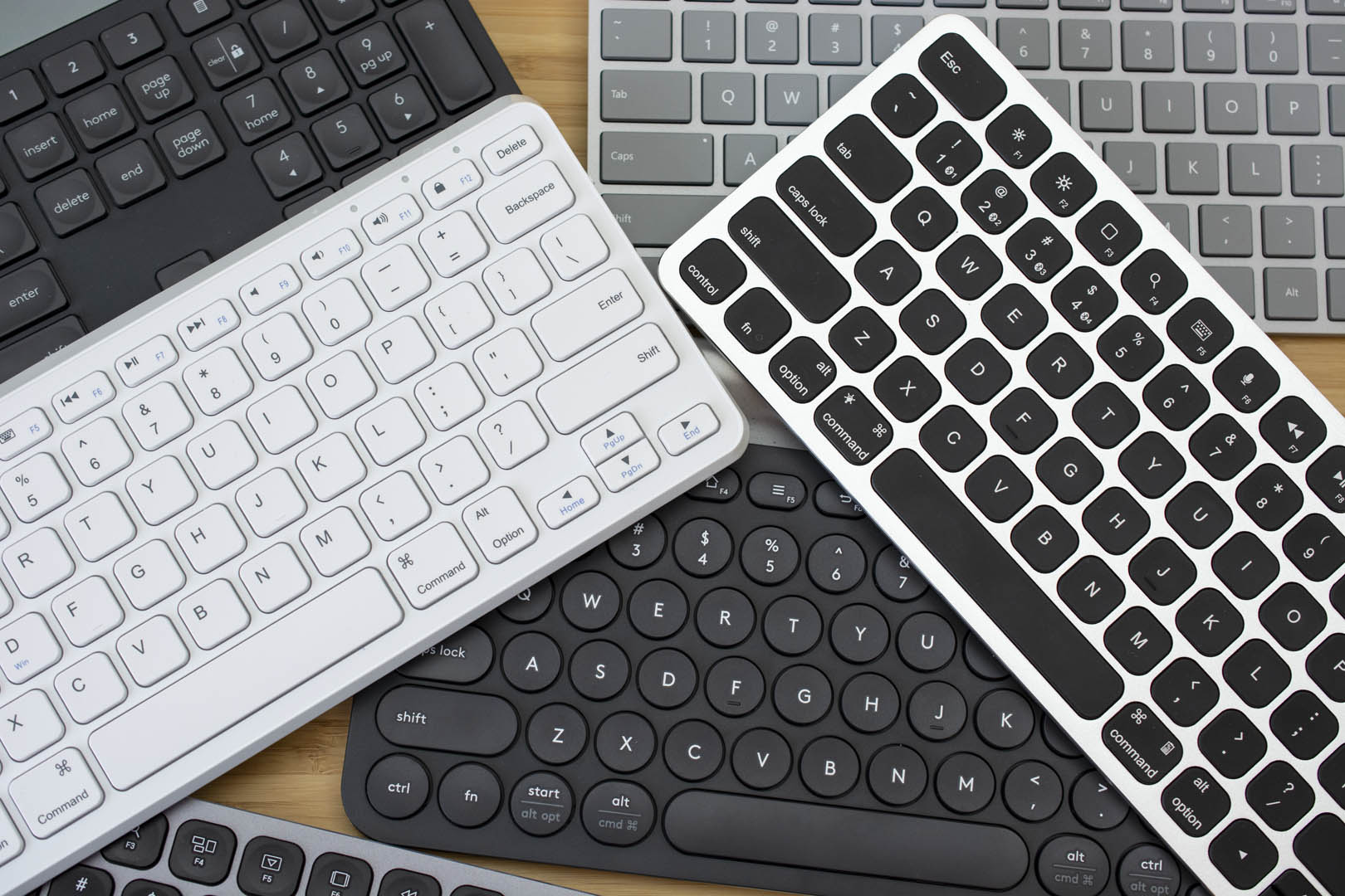 5 keyboards are stacked on top and beside each other. All of them are slightly different from one another with various key shapes and sizes. Colours are as follows: All black, all white, grey, silver with black keys.