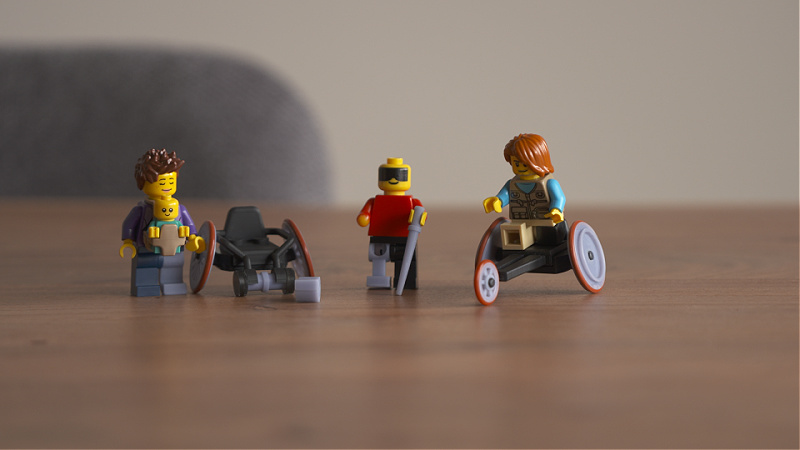 Lego people sit on a table top. One is seated in a wheelchair. Another is holding a cane.
