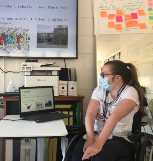 A young woman sits at a desk, looking at a laptop. She is wearing a medical mask.