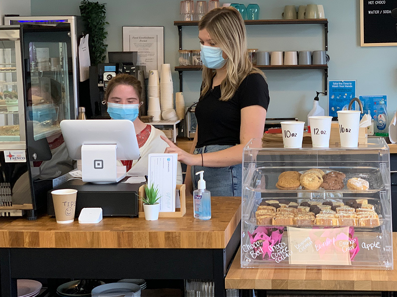 Two women, wearing surgical masks, stand behind a cash register. A case full of baked goods is to their left.