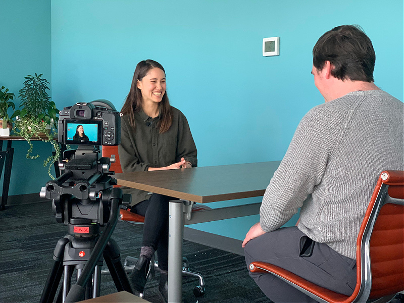 A woman, seated at a table, laughs while being filmed for a TV segment.