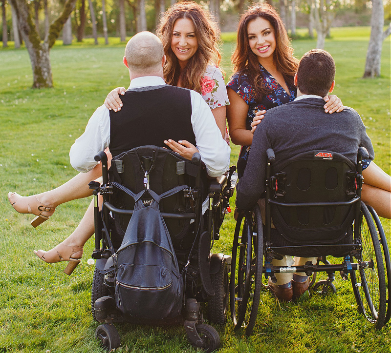 Two women, sitting in the laps of two male wheelchair users, smile into the camera.