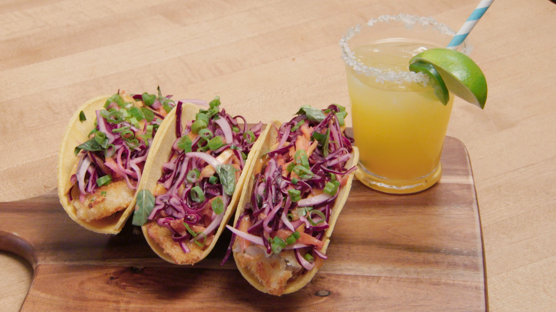 A trio of tacos sit on a table. A mocktail sits next to them.