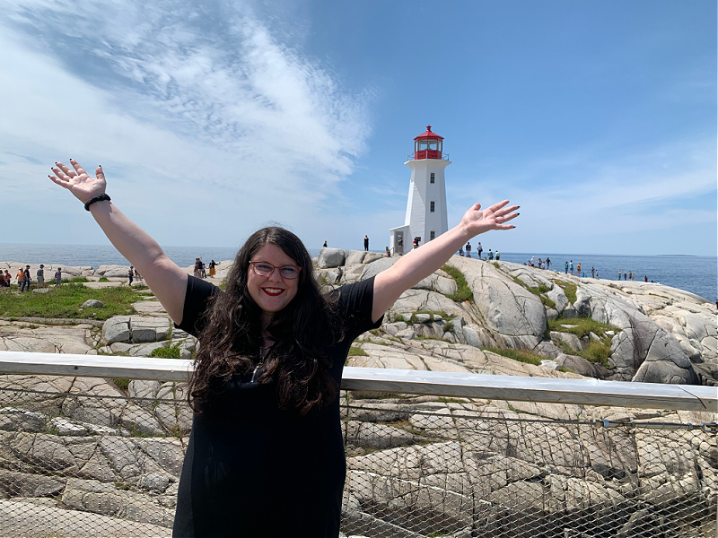 Jillian Gillis smiles into the camera, holding her arms above her head. The Peggy's Cove lighthouse is in the background.