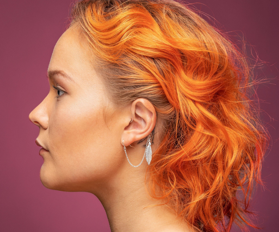 A woman is shown wearing a Deafmetal feather accessory on her hearing aid.