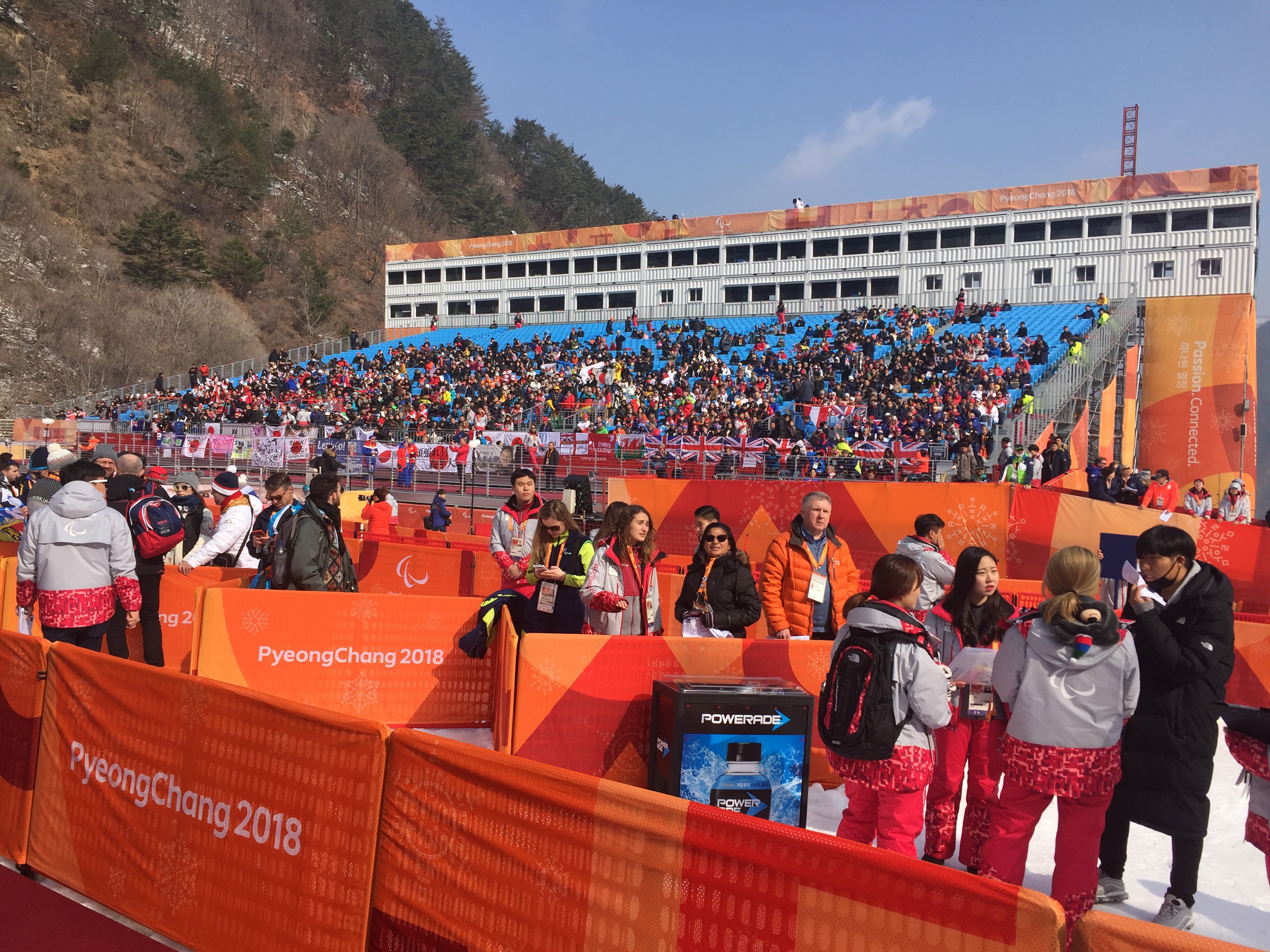 View of the grandstands. A packed crowd made up of countries from around the world cheers on their respective athletes. Many flags from different nations are draped over the bottom railing. 