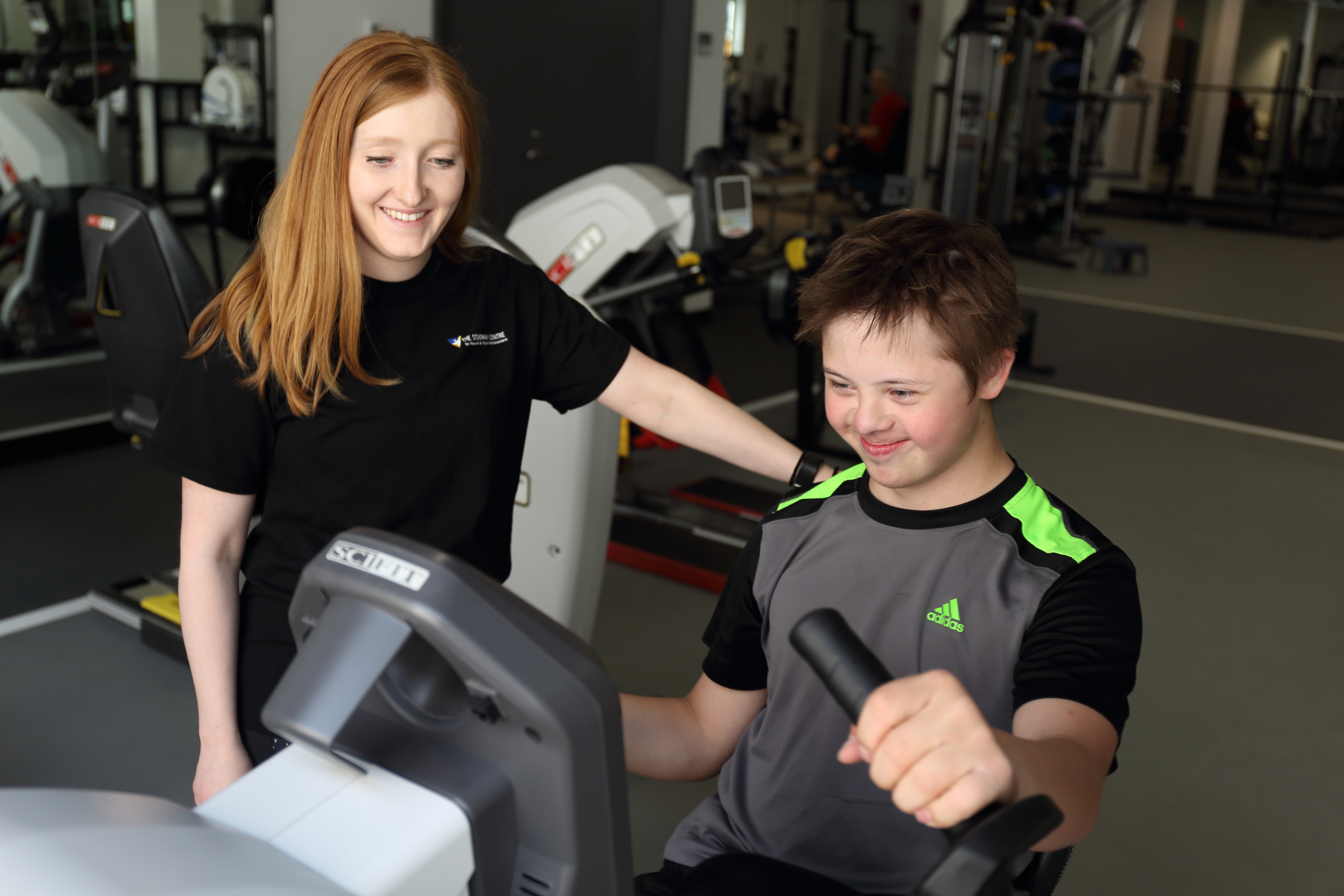 A female coach watches over a boy who is using a piece of exercise equipment.