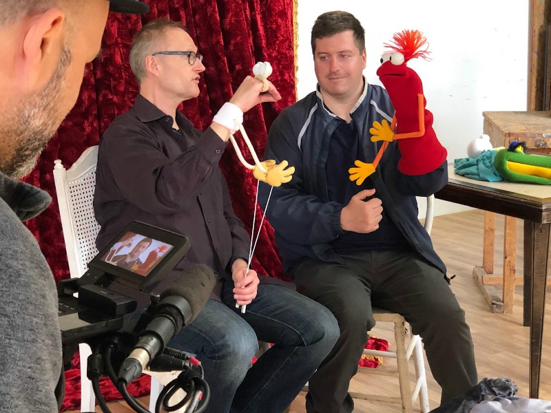 Two men sit in chairs, holding puppets. A television camera films them.