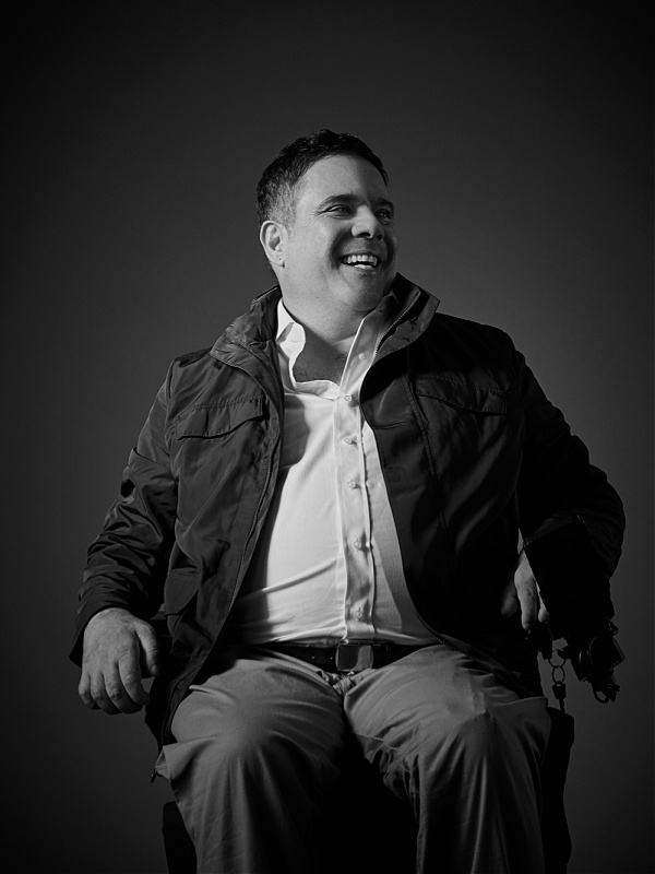 Chris smiles into the camera. He is seated in a power wheelchair.
