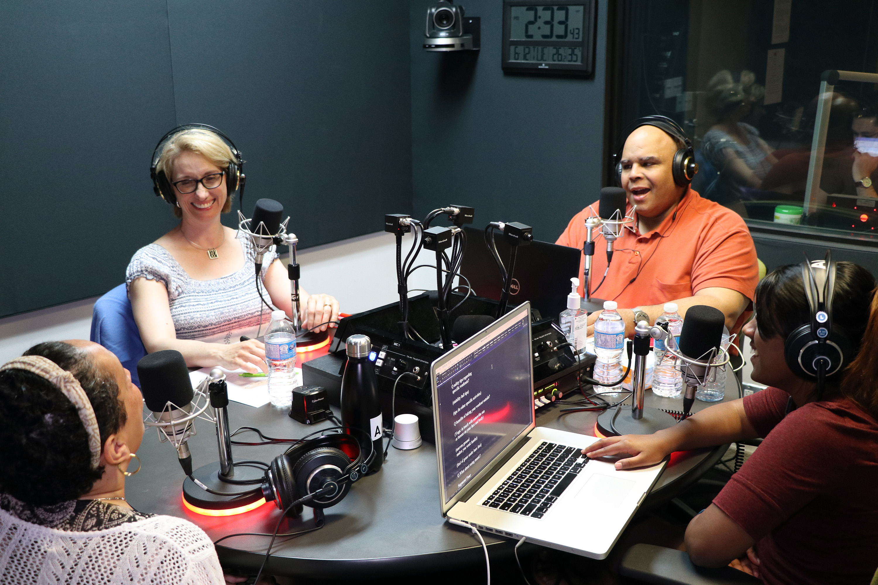 Kim sits around the table in studio with Kelly, Ramya and a guest. They are all wearing headphones and she looks on at the guest smiling.