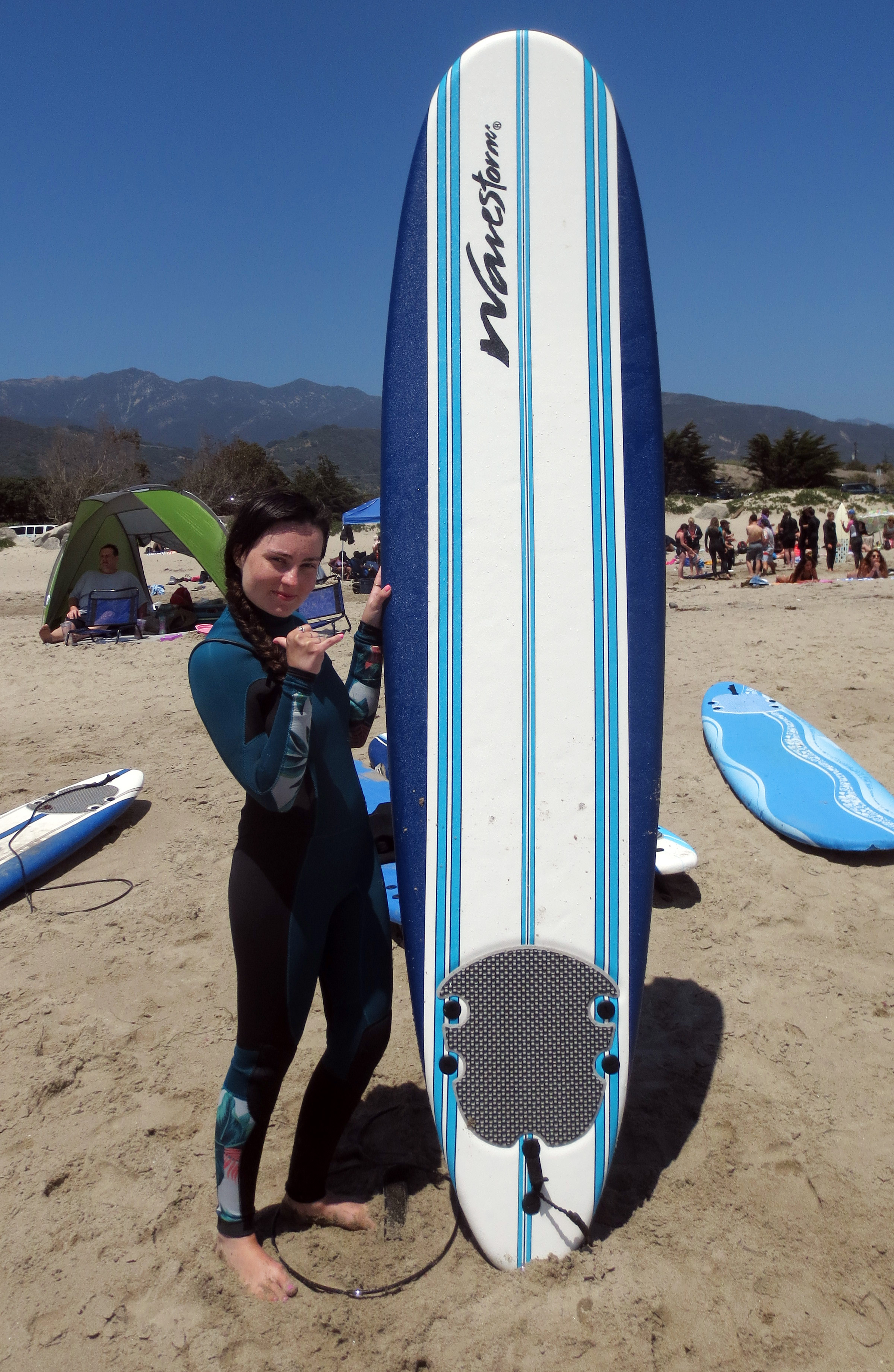 AMI This Week’s Molly Burke holds her surfboard on a sunny California beach wearing a stylish Billabong wetsuit. The surfboard is much taller than she is.