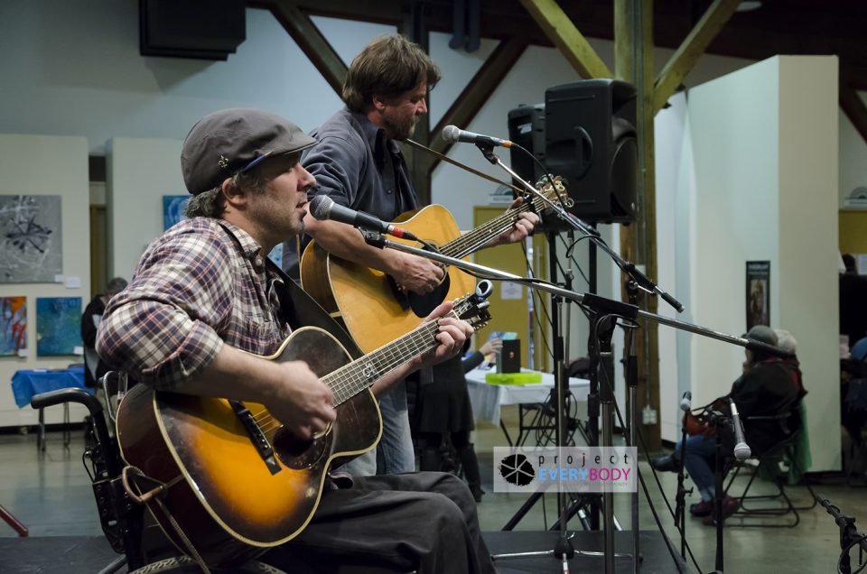 Two men play guitar and sign on stage. They are seated. The Project EveryBODY logo is in the corner of the picture.