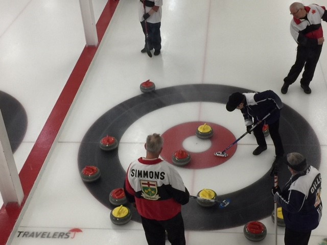 Curlers from a past tournament are on the ice. Several athletes look on as a woman uses a broom to propel a rock forward.