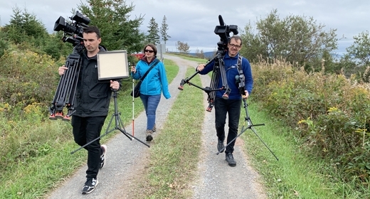 Three people walk along a trail. They are carrying television cameras.